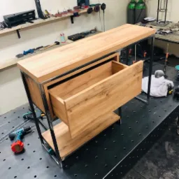 Biurko z blatem i szufladą z drewna w odcieniach brązu, osadzone na metalowym stelażu, widoczne w warsztacie na perforowanym stole spawalniczym, z wkrętarką obok.