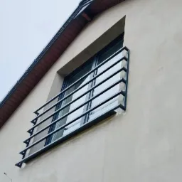 Zabezpieczenie okna na poddaszu wykonane z czarnego metalu, z poziomymi prętami i trójkątnymi wspornikami, zamontowane na jasnej elewacji budynku.