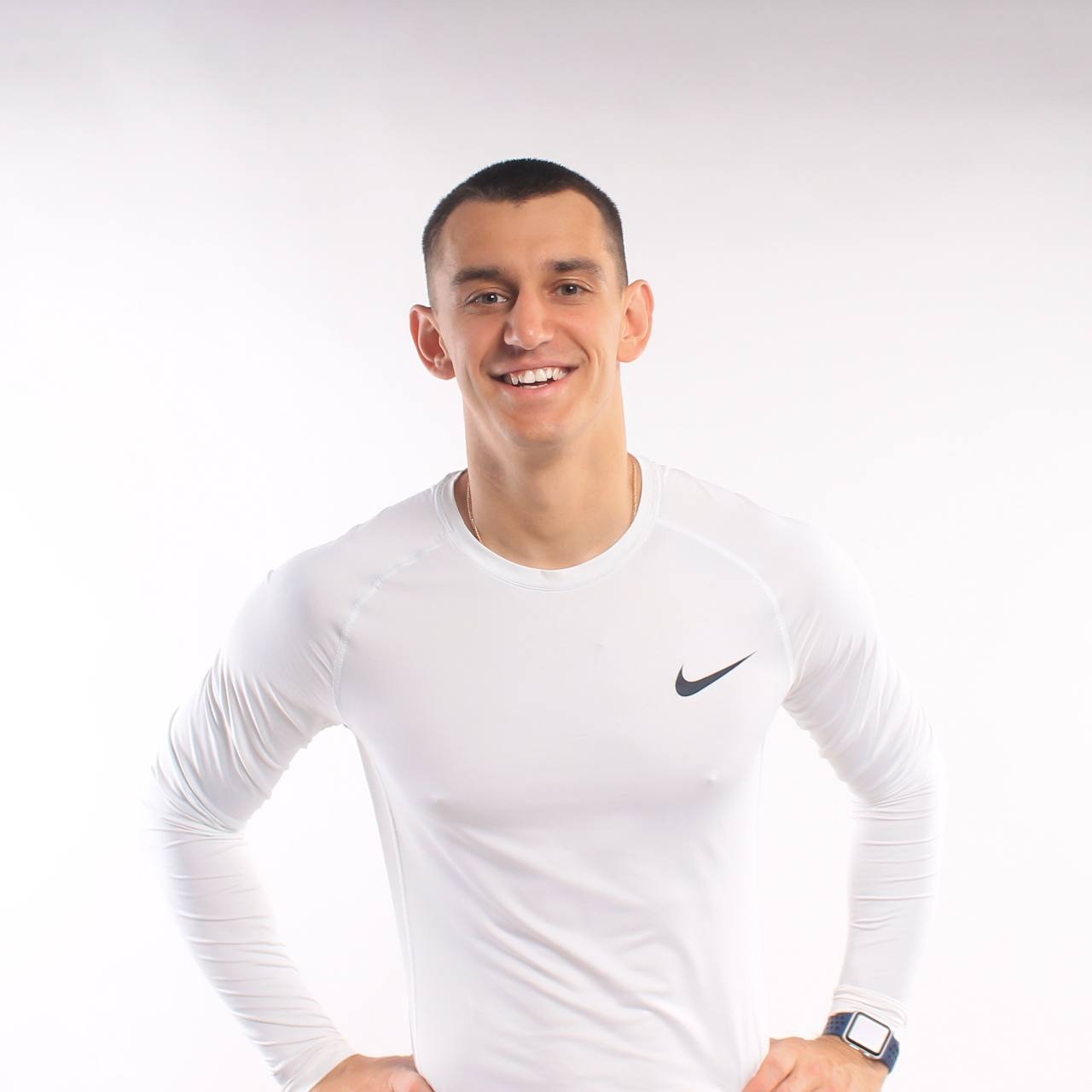 Uśmiechnięty mężczyzna w białej koszulce z długim rękawem i logo, pozujący na białym tle, z zegarkiem na nadgarstku.
