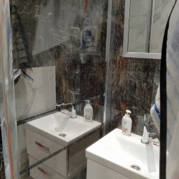 Stefan Olszynka - Wykończona łazienka z kabiną prysznicową i dwiema umywalkami z szafkami. Ściany wyłożone płytkami imitującymi marmur. Widoczne detale armatury i kosmetyki.