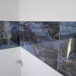 Stefan Olszynka - Fragment ściany wykończonej marmurowymi płytkami w odcieniach szarości i błękitu, z widocznymi puszkami elektrycznymi w rogu i fragmentem drewnianej podłogi.