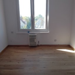 Stefan Olszynka - Jasne panele podłogowe w pokoju z oknem i grzejnikiem. Widoczne listwy przypodłogowe i gniazdka elektryczne. Naturalne światło dzienne.