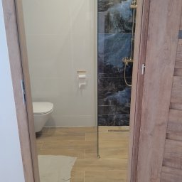 Stefan Olszynka - Nowoczesna łazienka z kabiną typu walk-in, wykończona płytkami imitującymi marmur i drewnopodobną podłogą. Widok przez uchylone drzwi.