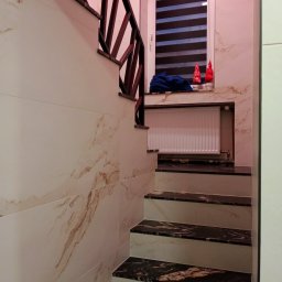 Stefan Olszynka - Wykończone schody z marmurowymi stopniami i drewnianą balustradą, ściany wyłożone płytkami imitującymi marmur, okno z roletą w tle. Dekoracje świąteczne na parapecie.