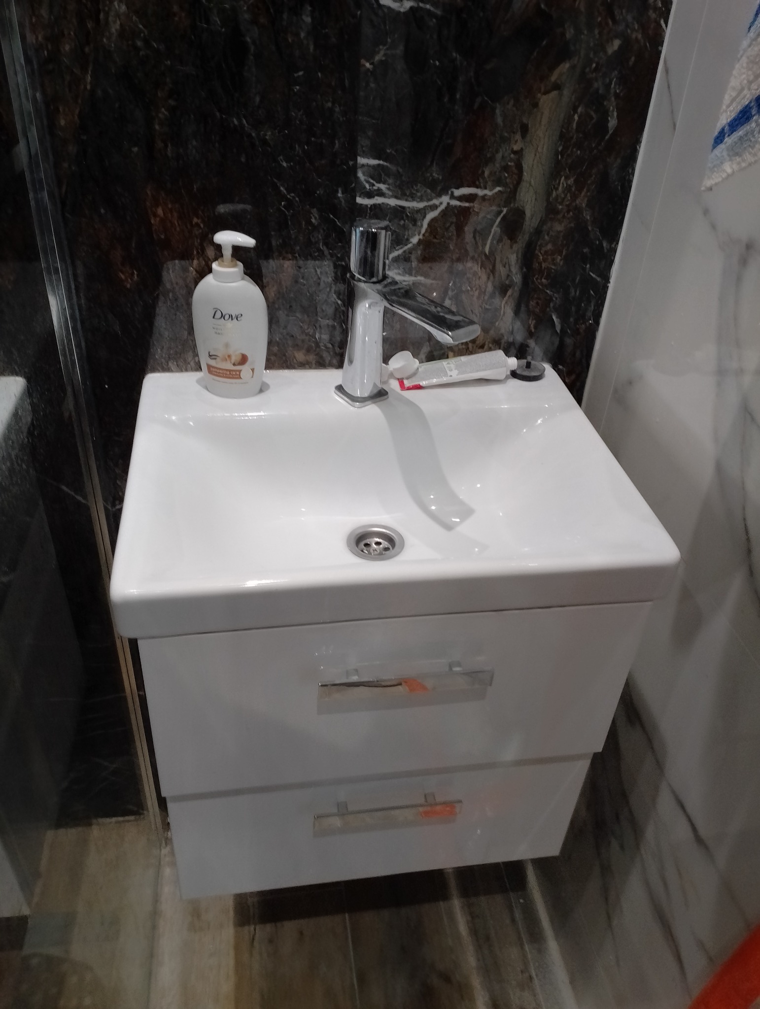 Biała umywalka z szafką w łazience z marmurową ścianą. Bateria chromowana, dozownik mydła Dove i tuba pasty do zębów na blacie. Widoczne dwie szuflady z uchwytami.