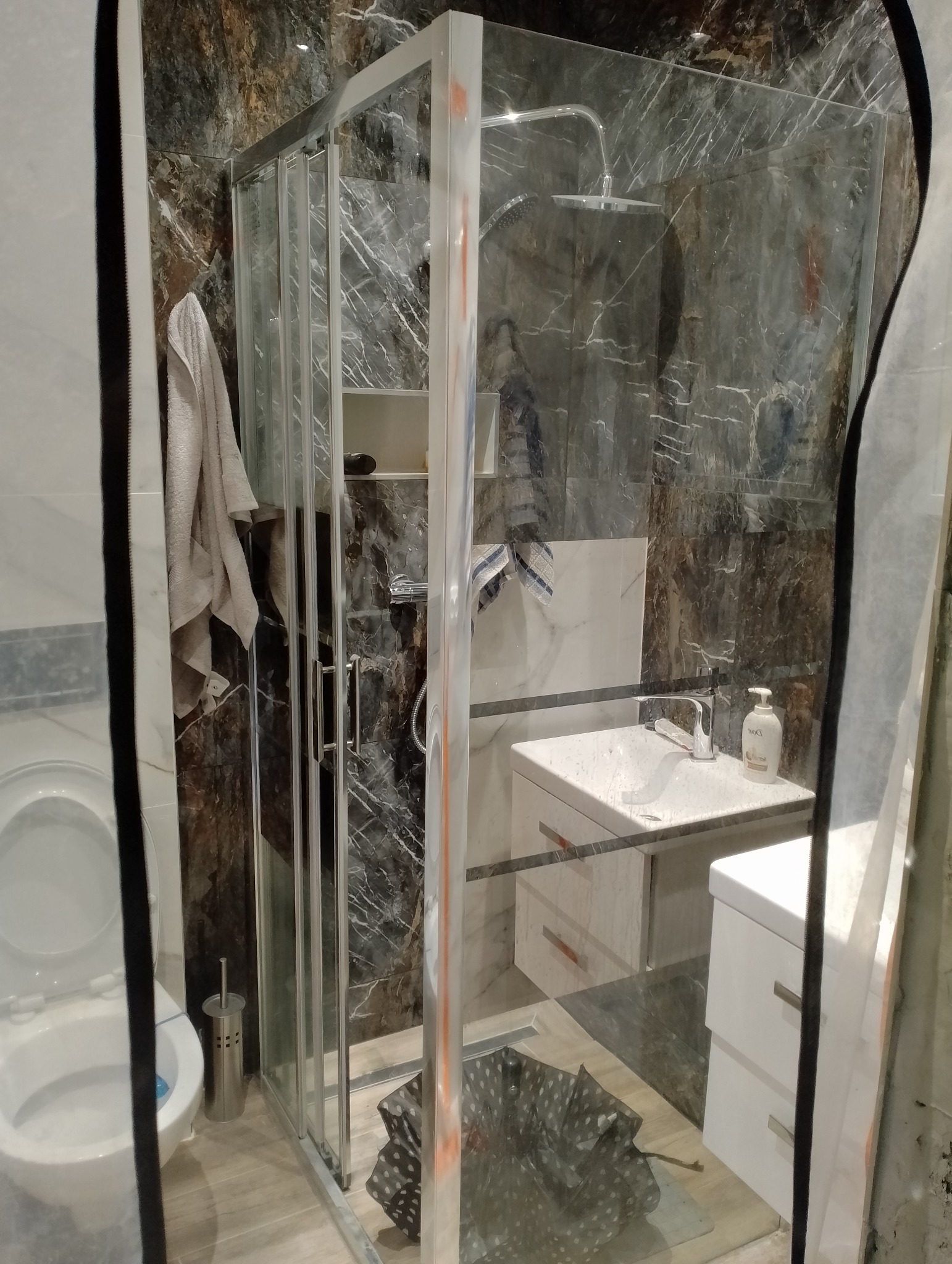 Nowoczesna łazienka z kabiną prysznicową typu walk-in, umywalką nablatową i toaletą. Ściany wykończone płytkami imitującymi marmur. Widoczny ręcznik i akcesoria.