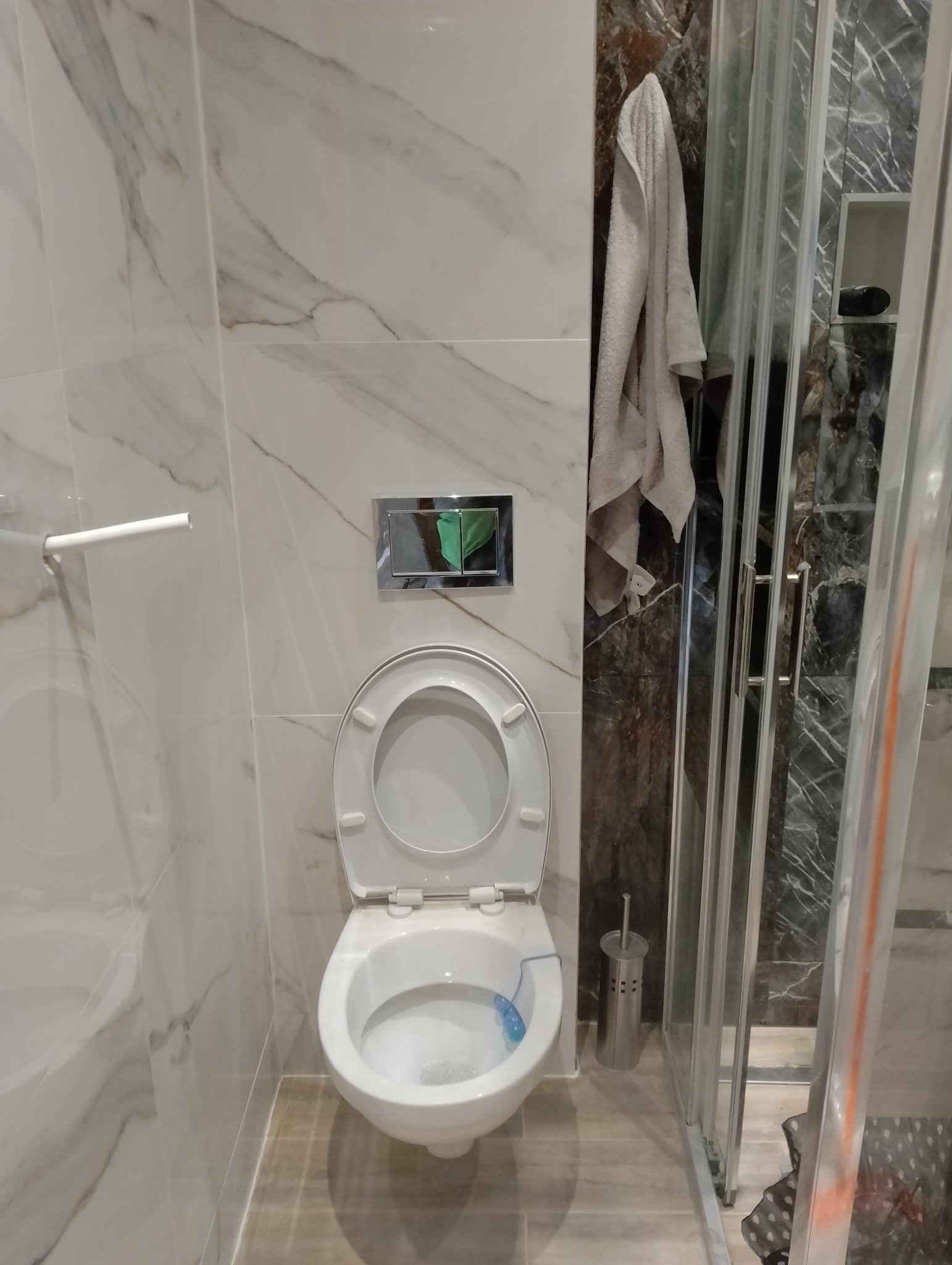 Nowoczesna łazienka z marmurowymi płytkami, kabiną prysznicową i podwieszaną toaletą z chromowanym przyciskiem spłukującym. Widoczny ręcznik i szczotka do WC.