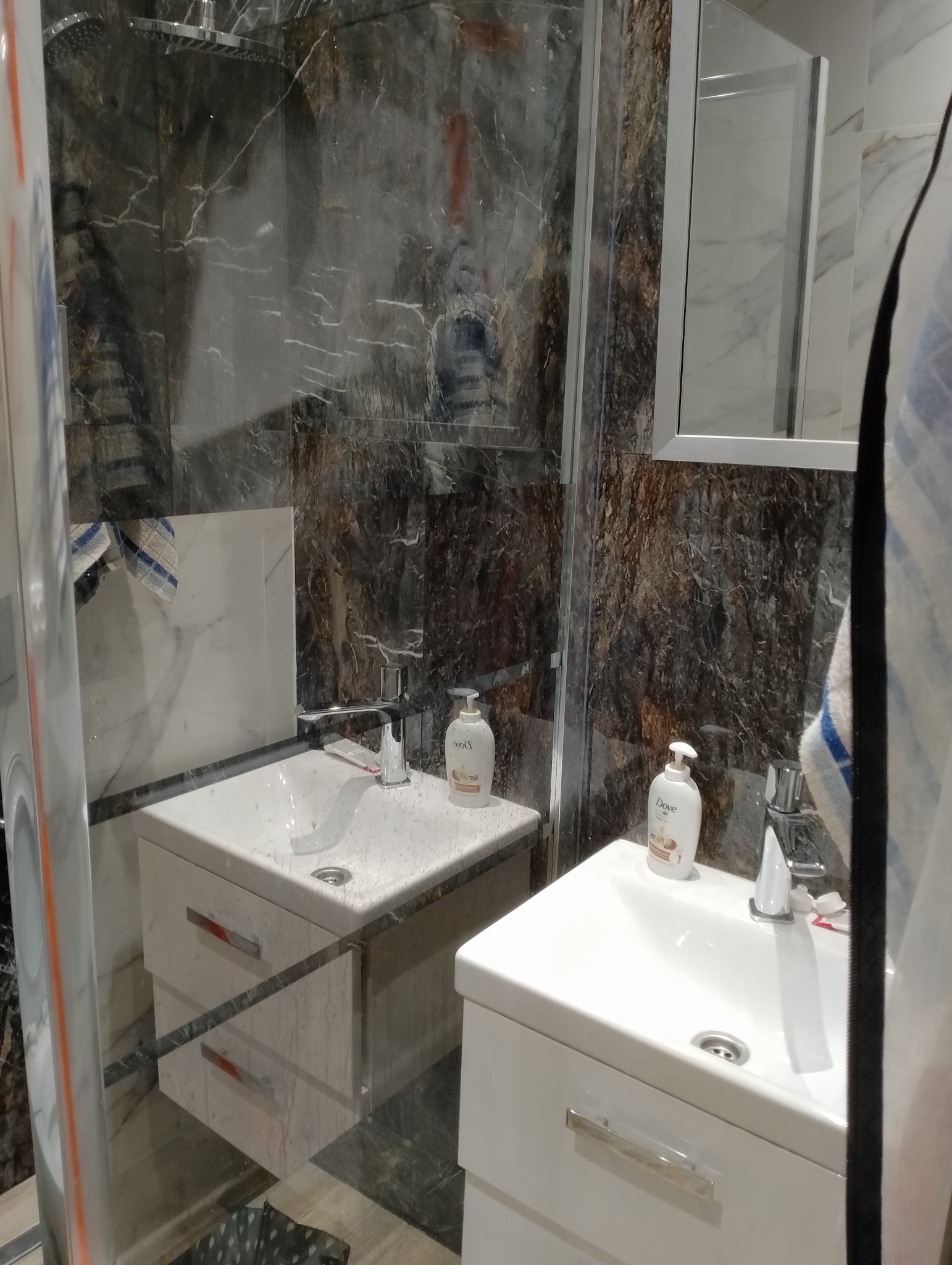 Wykończona łazienka z kabiną prysznicową i dwiema umywalkami z szafkami. Ściany wyłożone płytkami imitującymi marmur. Widoczne detale armatury i kosmetyki.