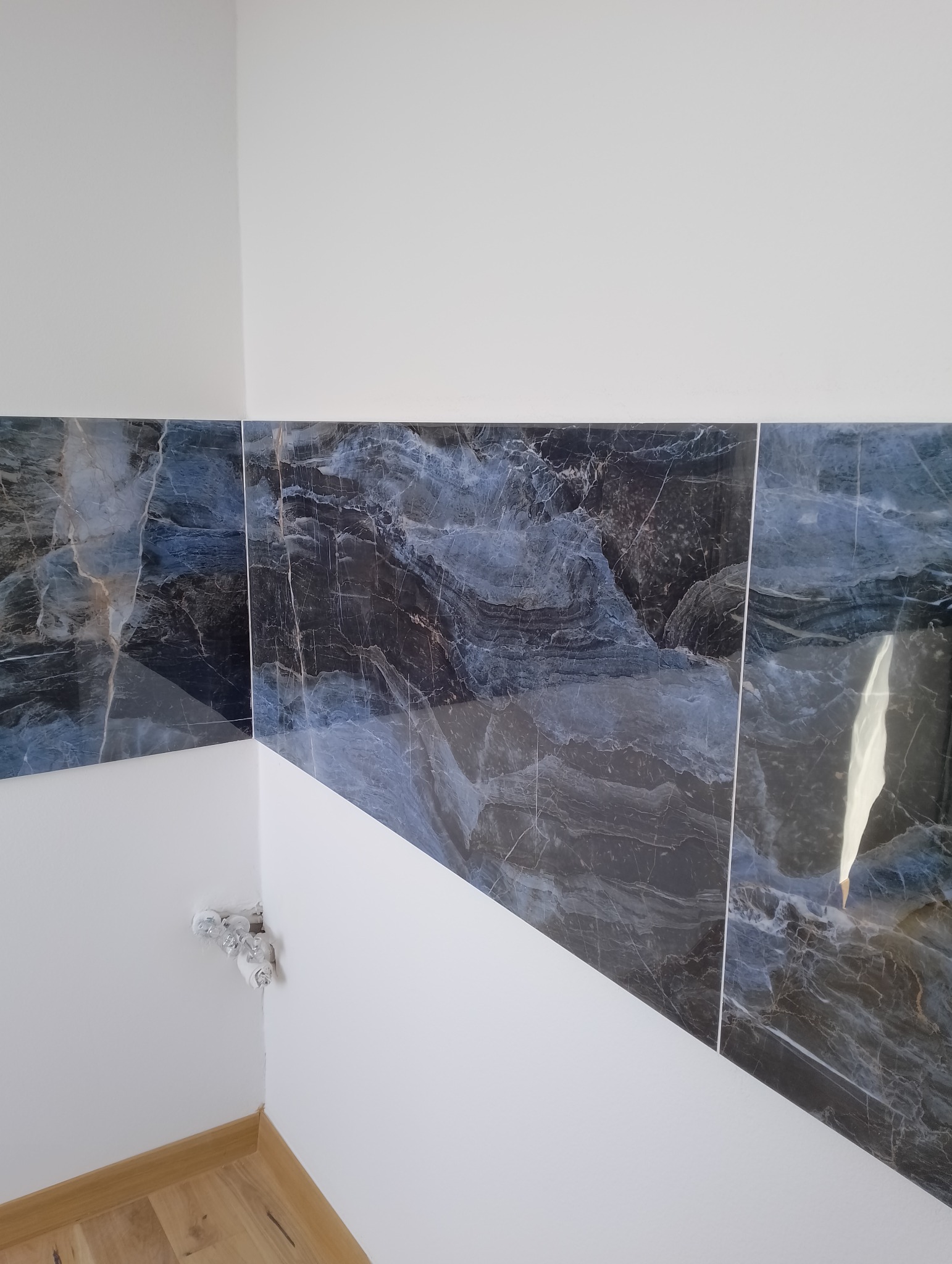 Fragment ściany wykończonej marmurowymi płytkami w odcieniach szarości i błękitu, z widocznymi puszkami elektrycznymi w rogu i fragmentem drewnianej podłogi.