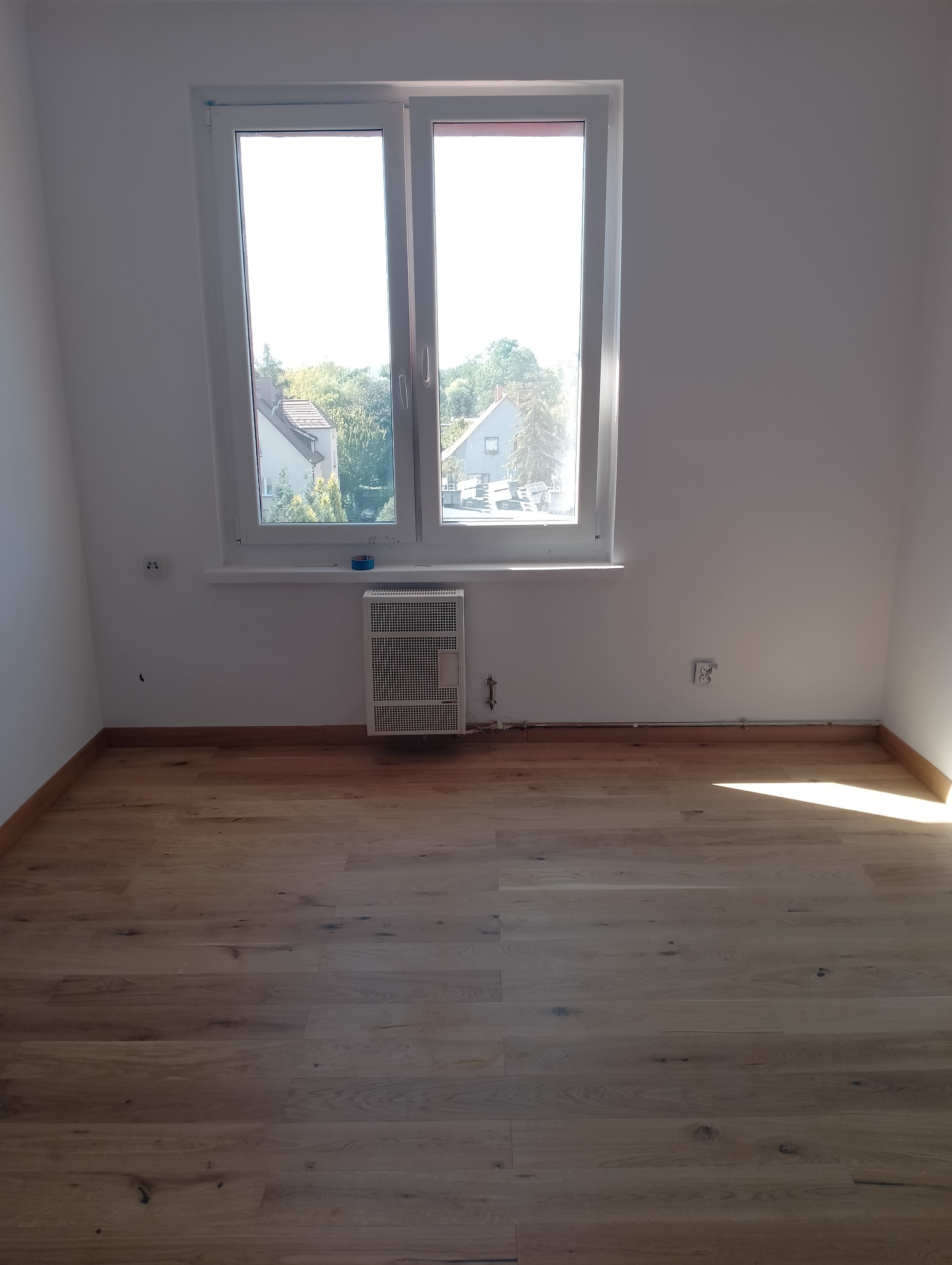 Jasne panele podłogowe w pokoju z oknem i grzejnikiem. Widoczne listwy przypodłogowe i gniazdka elektryczne. Naturalne światło dzienne.