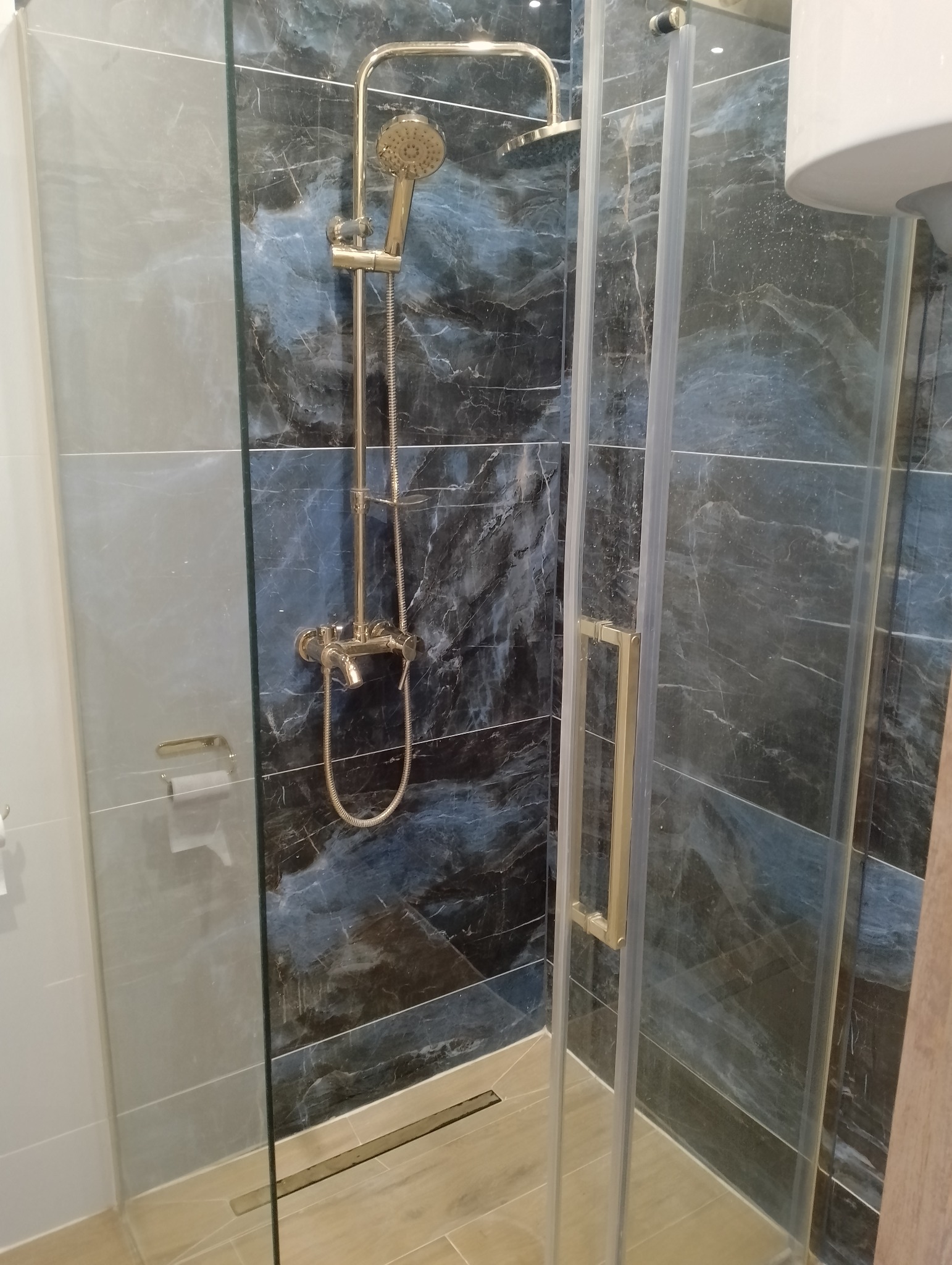 Kabina prysznicowa z deszczownicą w odcieniach złota i marmurowymi płytkami w kolorach szarości i granatu. Nowoczesny design z odpływem liniowym.