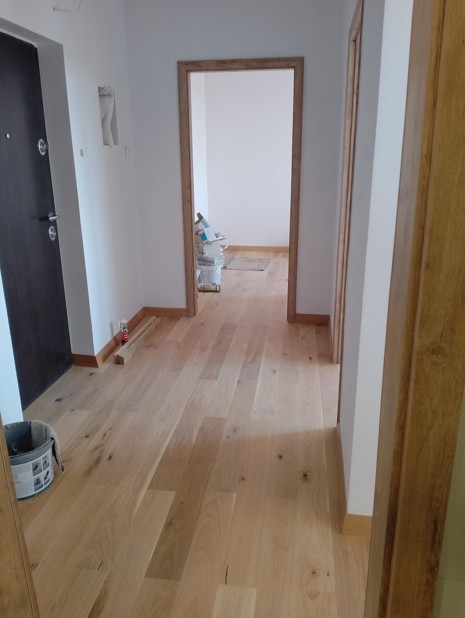 Jasne panele podłogowe ułożone w korytarzu z widocznymi listwami przypodłogowymi i ościeżnicami. W tle wiadro i materiały budowlane.