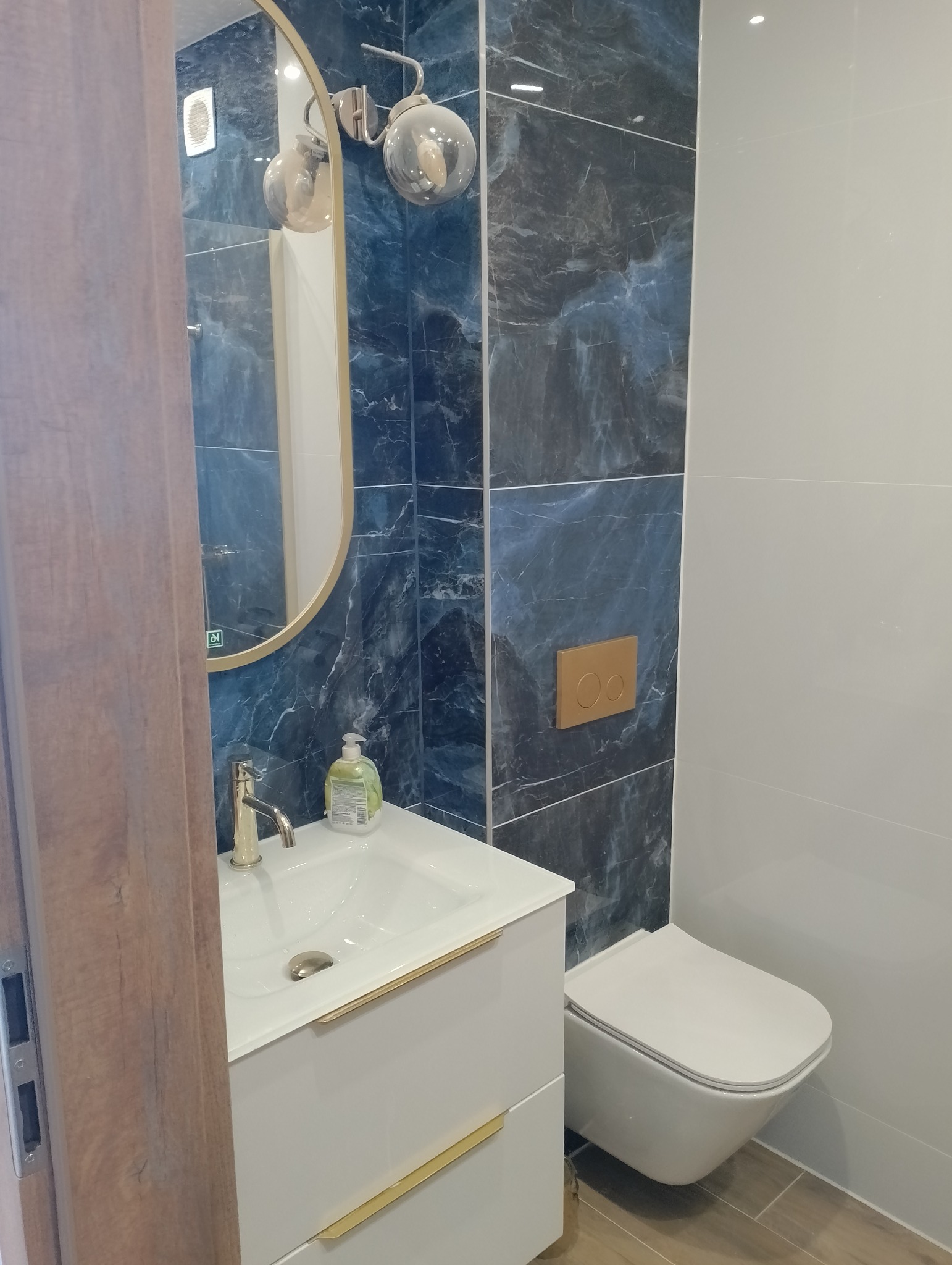 Nowoczesna łazienka z umywalką na szafce, toaletą podwieszaną i lustrem w złotej ramie. Ściany wyłożone płytkami w odcieniach bieli i granatu z marmurkowym wzorem.