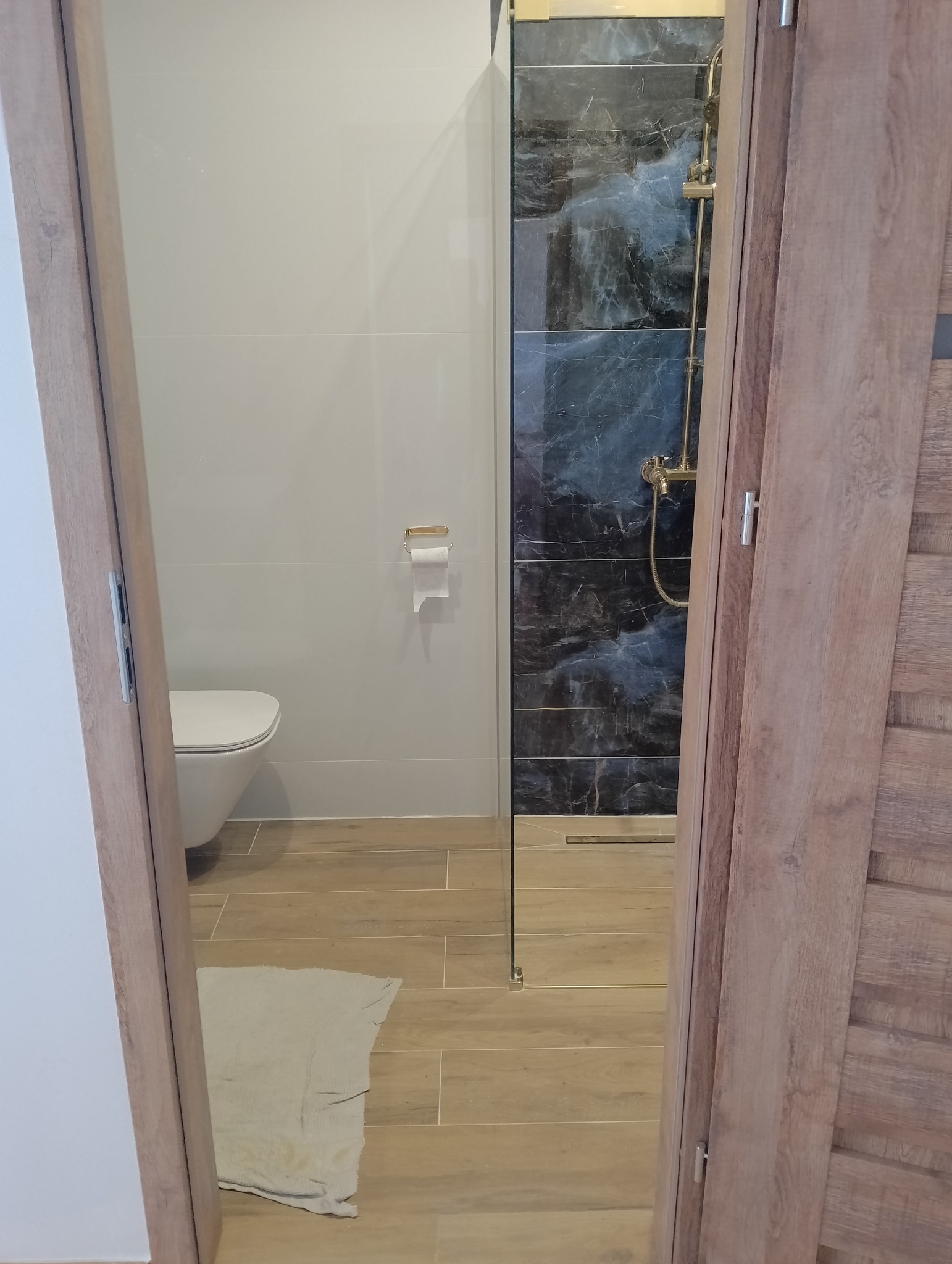 Nowoczesna łazienka z kabiną typu walk-in, wykończona płytkami imitującymi marmur i drewnopodobną podłogą. Widok przez uchylone drzwi.