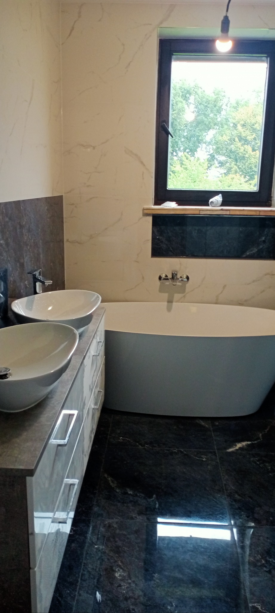 Nowoczesna łazienka z dwoma umywalkami na szafce z szufladami, białą wanną i oknem z widokiem na zieleń. Marmurowe płytki na ścianach i podłodze.