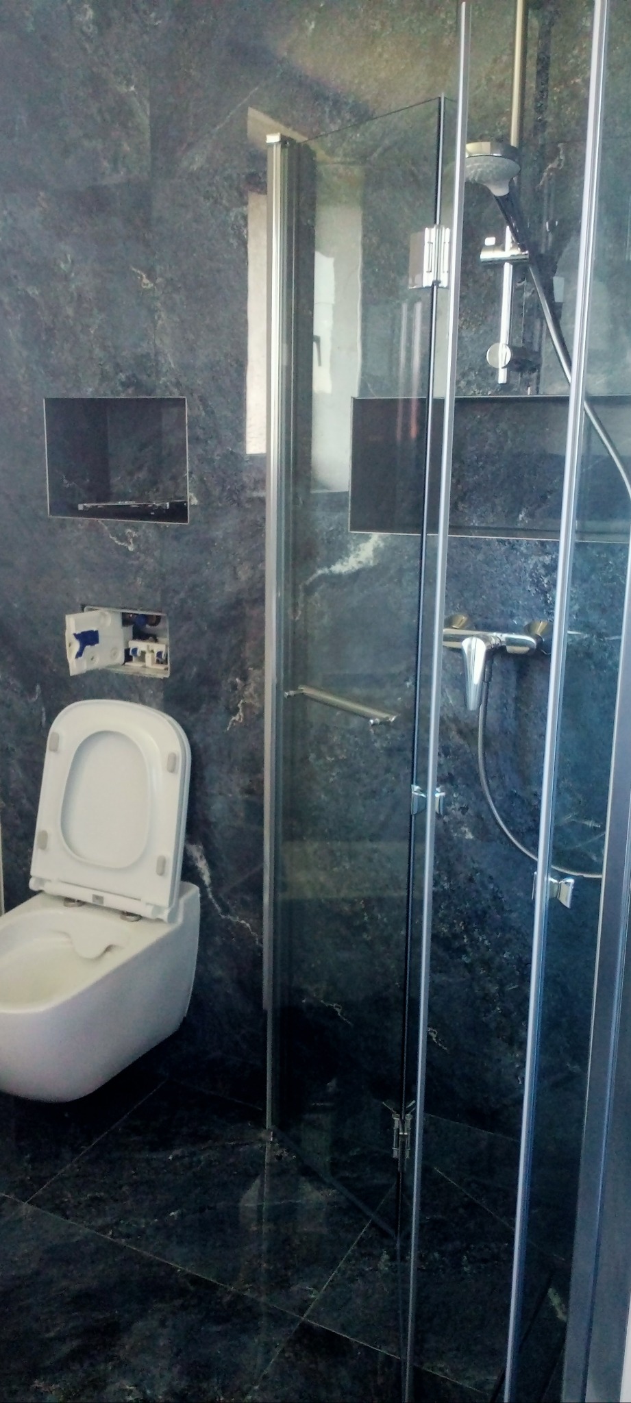 Nowoczesna łazienka z kabiną prysznicową i toaletą w zabudowie, wykończona ciemnym marmurem z wnękami. Widoczna instalacja podtynkowa.