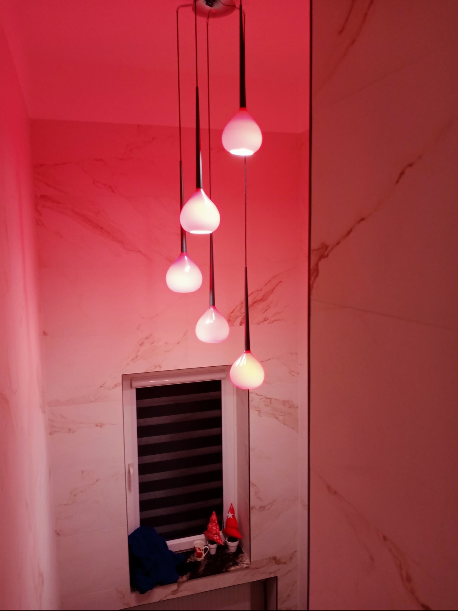 Nowoczesna łazienka z marmurowymi płytkami i designerską lampą wiszącą z pięcioma kulistymi kloszami, oświetlona czerwonym światłem. Widok na okno z żaluzjami.