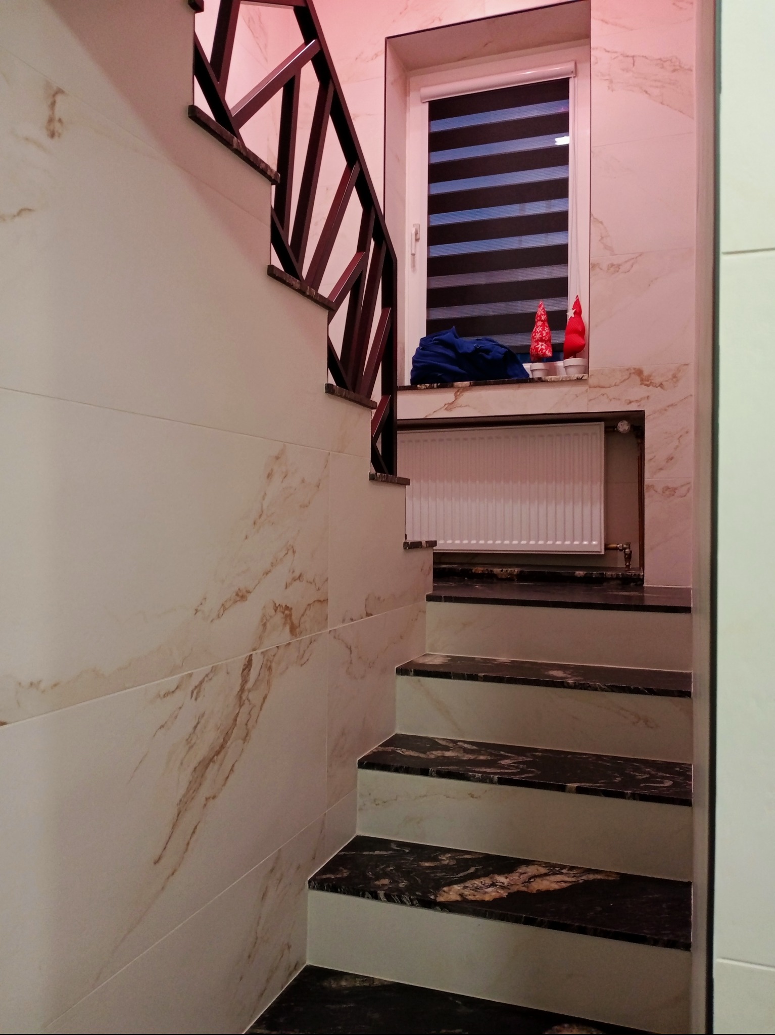 Wykończone schody z marmurowymi stopniami i drewnianą balustradą, ściany wyłożone płytkami imitującymi marmur, okno z roletą w tle. Dekoracje świąteczne na parapecie.