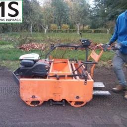 Mężczyzna obsługuje pomarańczową maszynę do układania trawy z rolki na przygotowanym podłożu w ogrodzie.