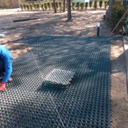 Pracownik w niebieskiej bluzie i czerwonych rękawicach układa plastikowe panele ażurowe wypełnione ziemią na piaszczystym podłożu, widoczne wytyczone sznurki i elementy oświetlenia ogrodowego w tle.