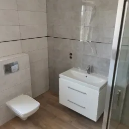 Nowoczesna łazienka z szarymi płytkami i białymi meblami: podwieszana toaleta z panelem, umywalka z szafką i kabina prysznicowa ze srebrnymi profilami.