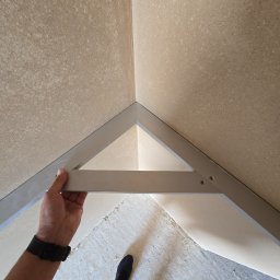 ASPEKT IAROSLAV BABII - Montaż metalowego profilu w rogu pomieszczenia z surowymi ścianami. Widoczna dłoń montera i fragment podłogi. Prace wykończeniowe w trakcie.