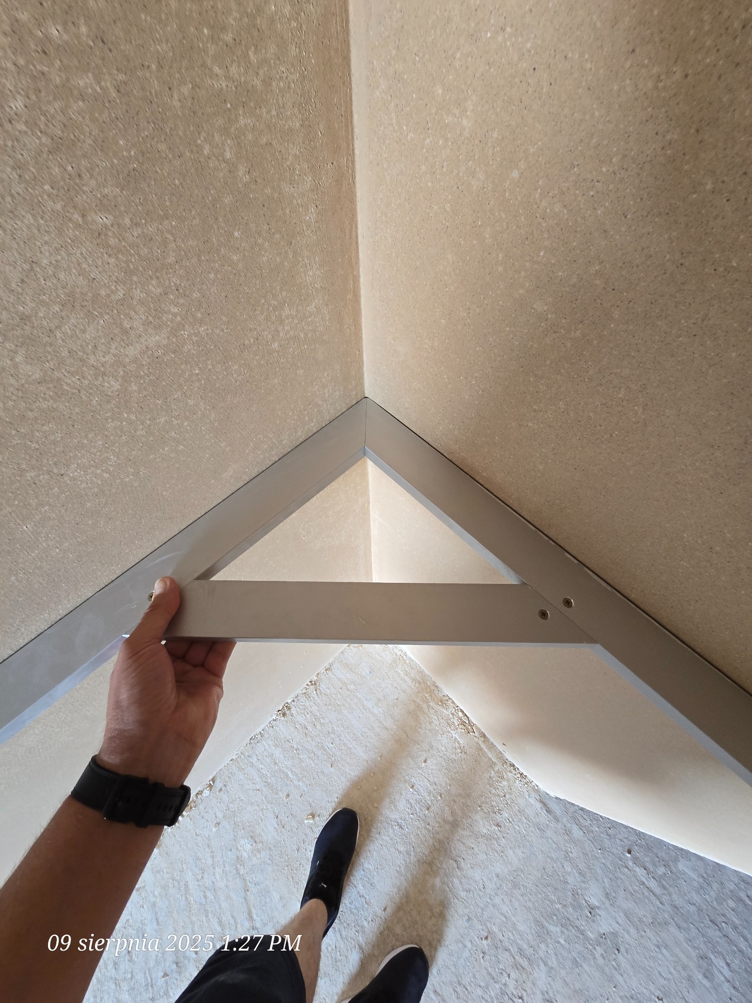 Montaż metalowego profilu w rogu pomieszczenia z surowymi ścianami. Widoczna dłoń montera i fragment podłogi. Prace wykończeniowe w trakcie.
