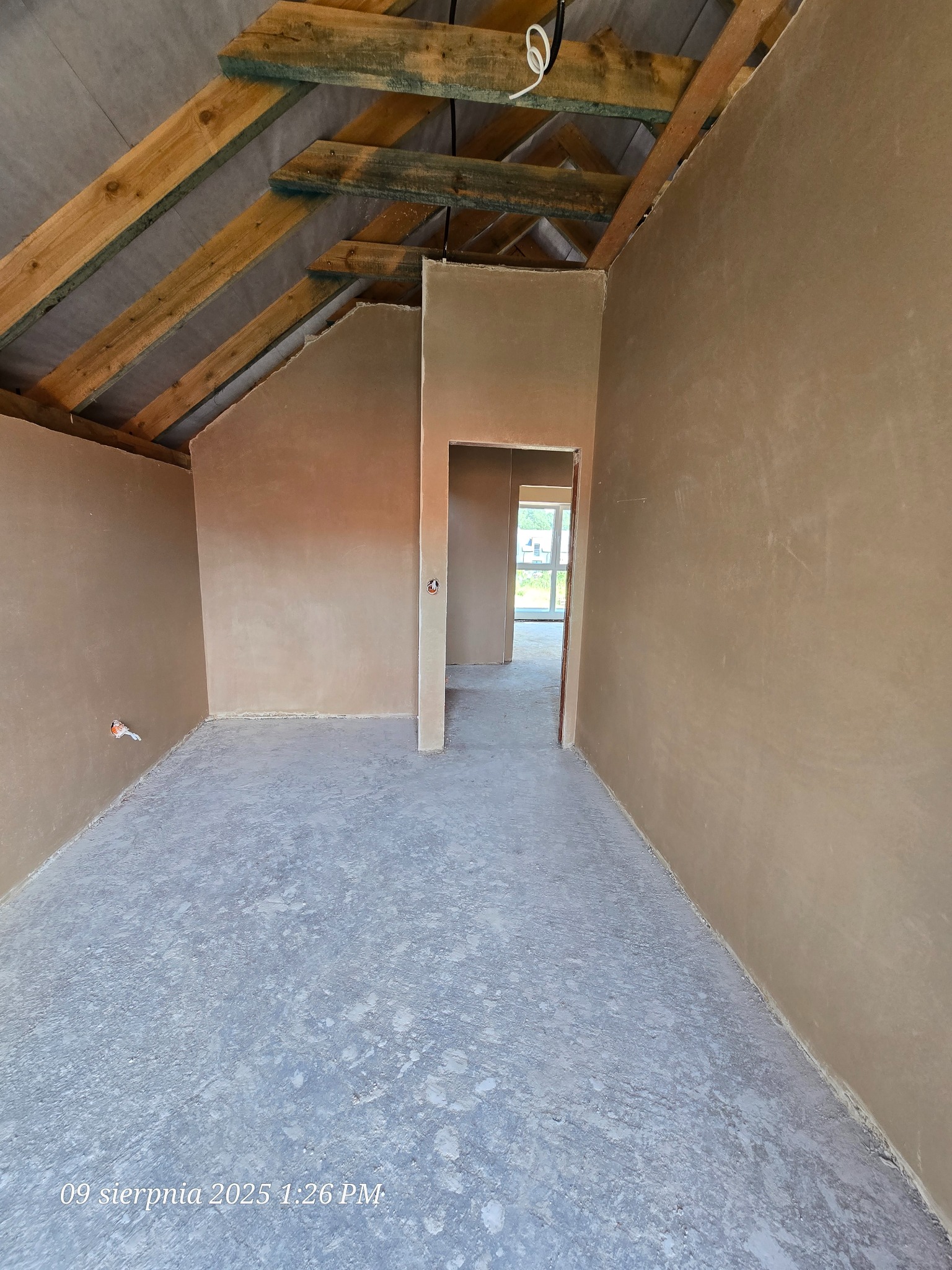 Surowe wnętrze poddasza z drewnianą konstrukcją dachu i świeżo nałożonymi tynkami na ścianach. Widoczne przygotowane otwory drzwiowe i surowy beton na podłodze.