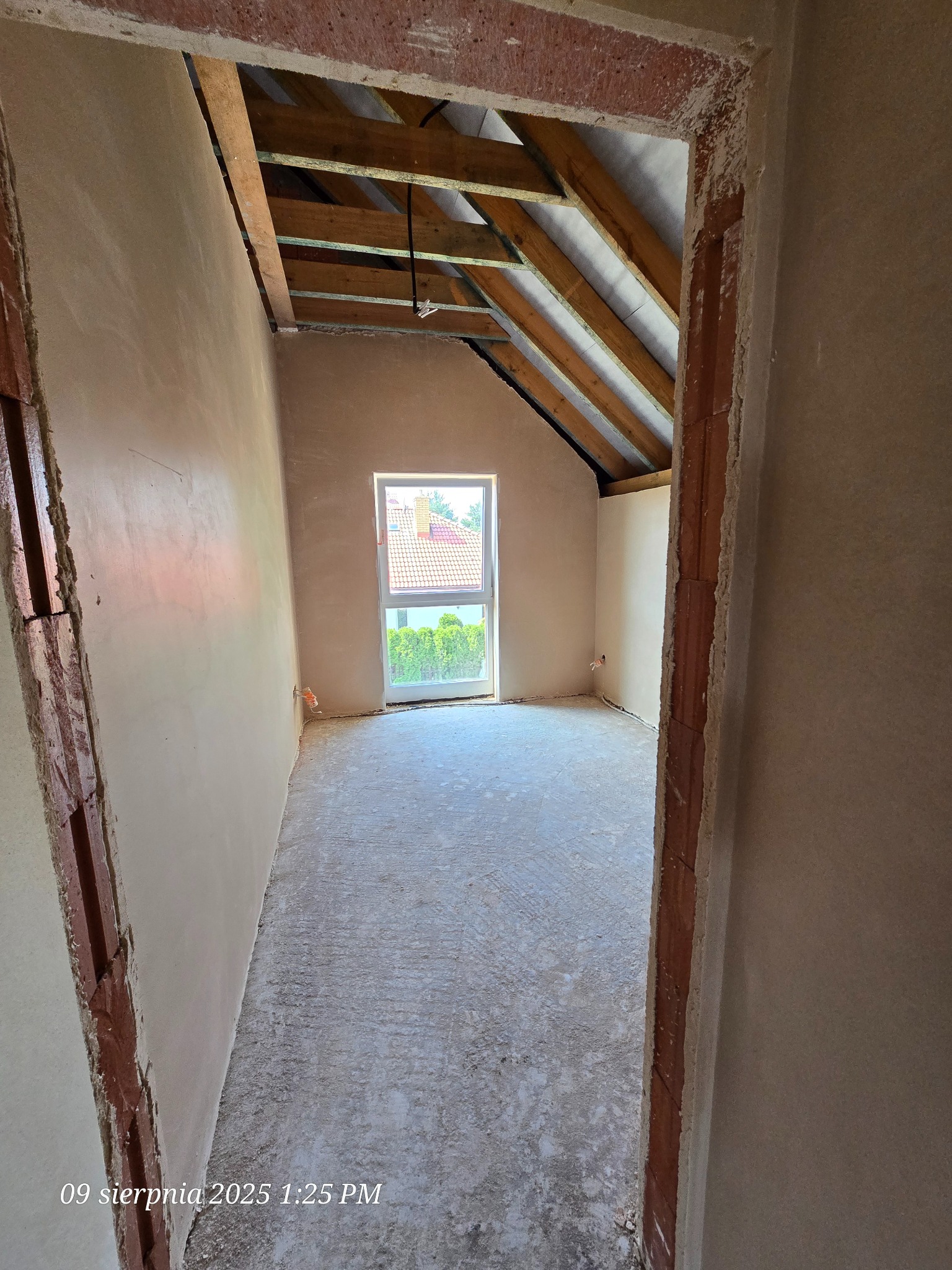 Surowe wnętrze poddasza po tynkowaniu, widok zza futryny. Ściany pokryte świeżym tynkiem, drewniana konstrukcja dachu, okno z widokiem na zieleń.
