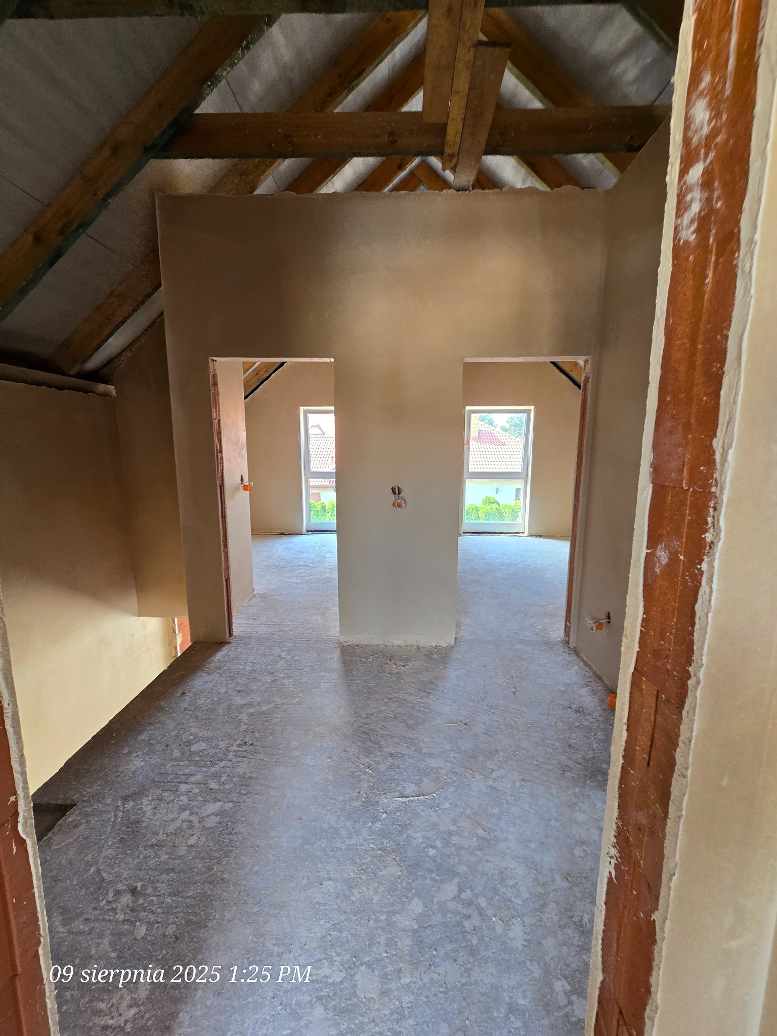 Wnętrze poddasza po tynkowaniu maszynowym, widoczne surowe drewniane belki stropowe i dwa symetryczne przejścia do kolejnych pomieszczeń z oknami na zewnątrz.