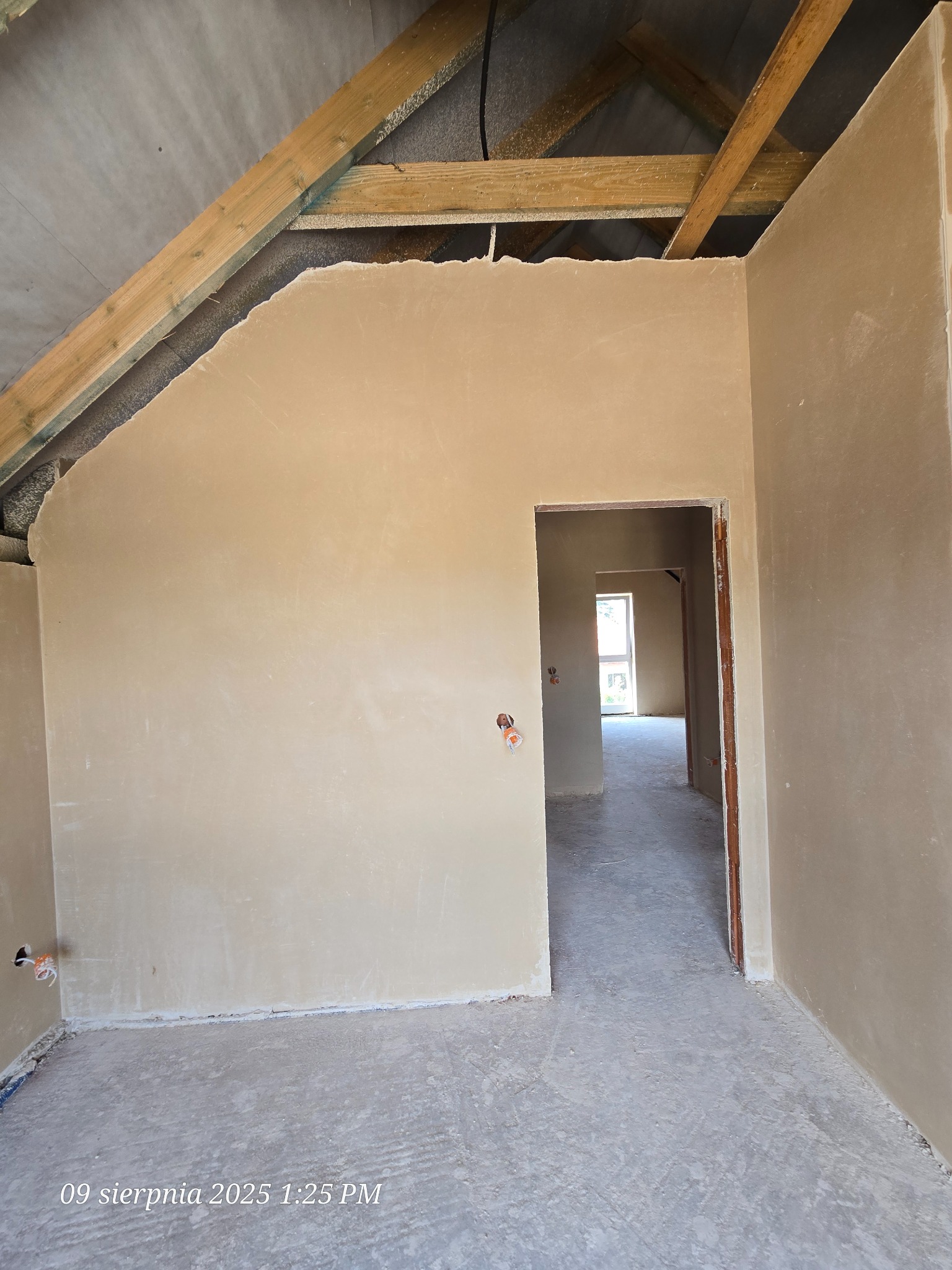 Wnętrze poddasza po tynkowaniu maszynowym, widoczne drewniane elementy konstrukcji dachu i gładkie ściany w odcieniach beżu. Otwór drzwiowy prowadzi do kolejnych pomieszczeń.