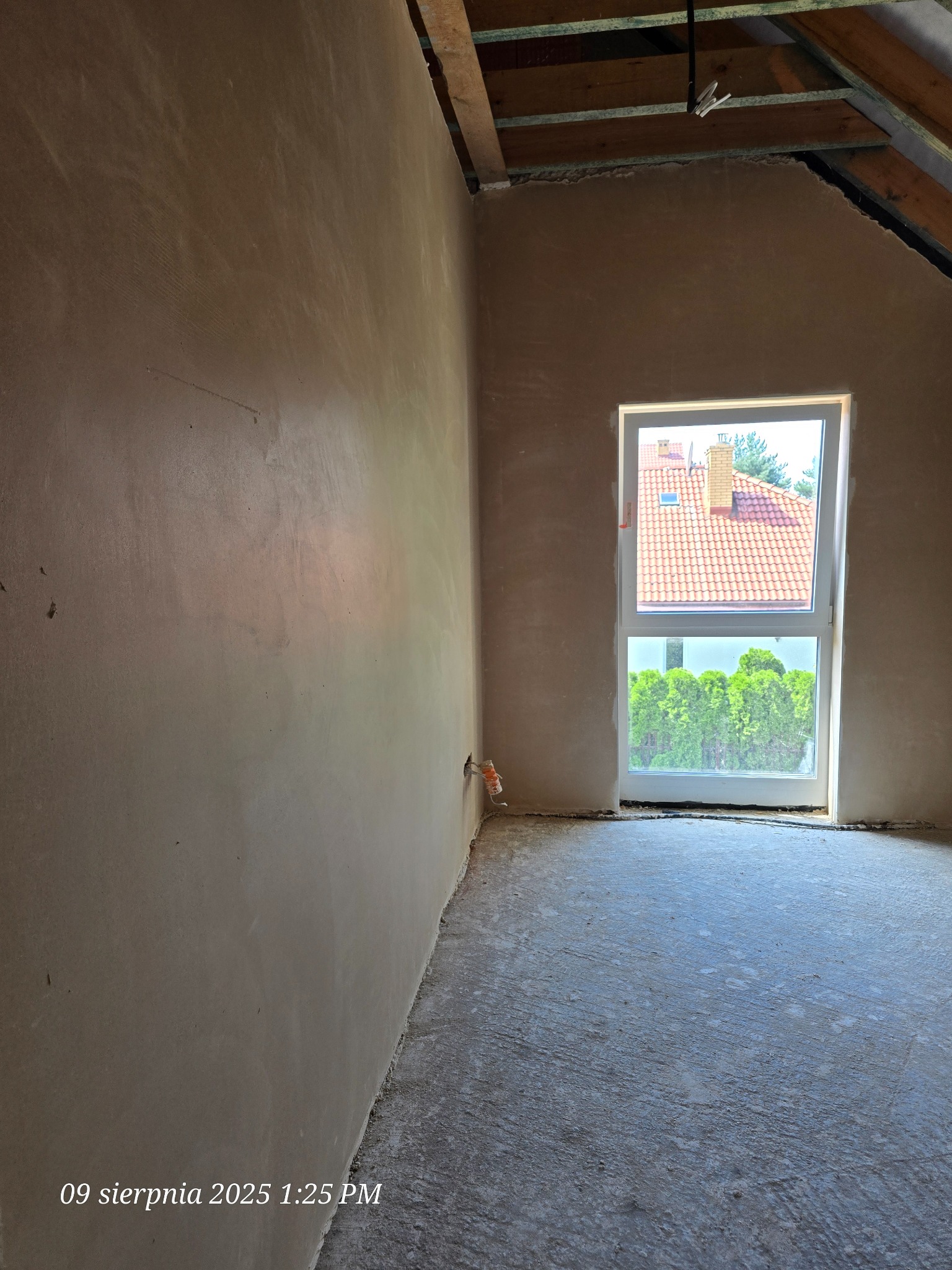 Pokój po tynkowaniu maszynowym, widok na ścianę z oknem. Surowy strop drewniany. Podłoże przygotowane pod wylewkę. Widok z okna na dom i zieleń.