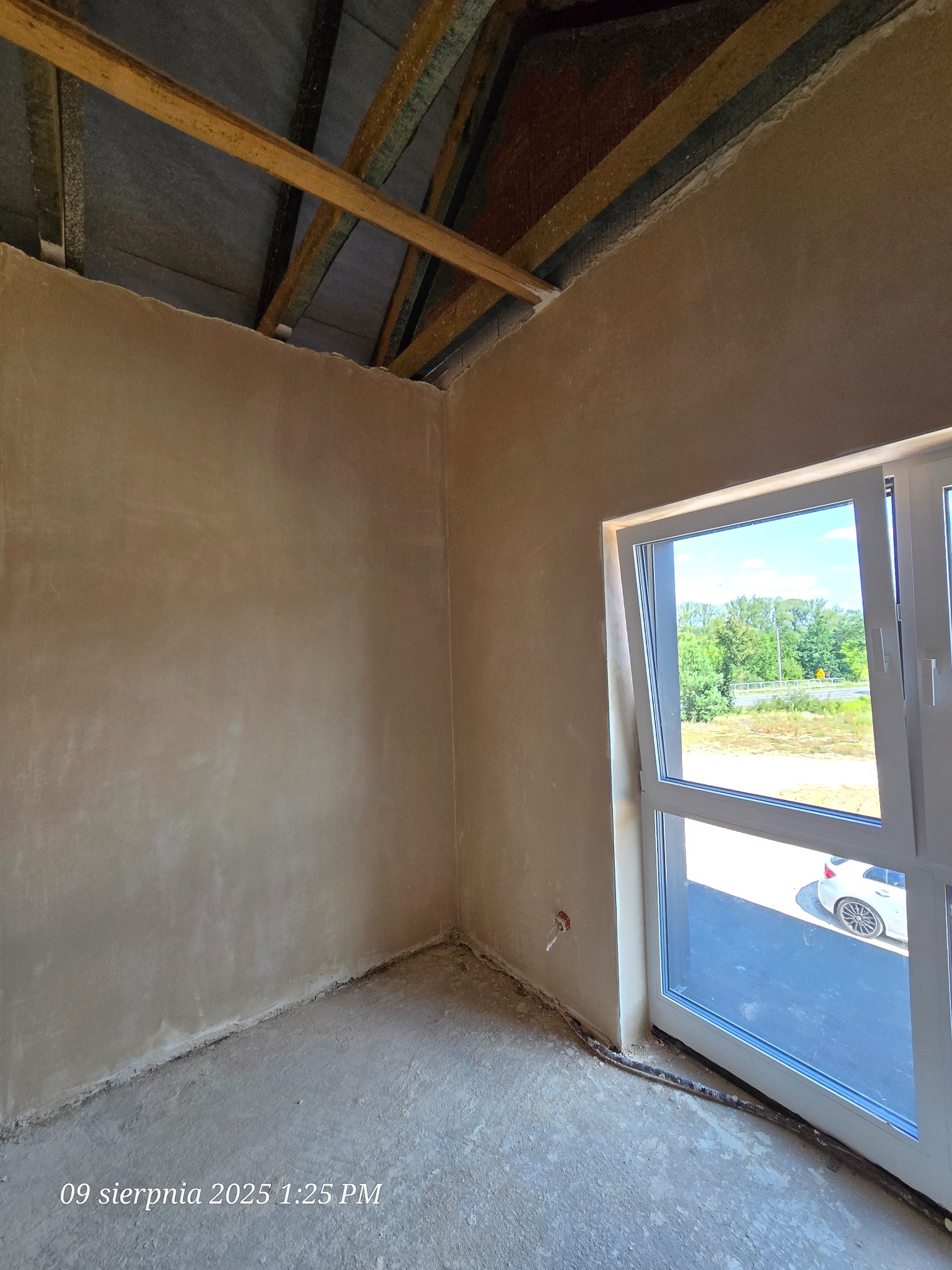 Wnętrze w stanie surowym: ściany po tynkowaniu, widoczne elementy konstrukcji dachu, okno z widokiem na zewnątrz. Instalacja elektryczna przy podłodze.