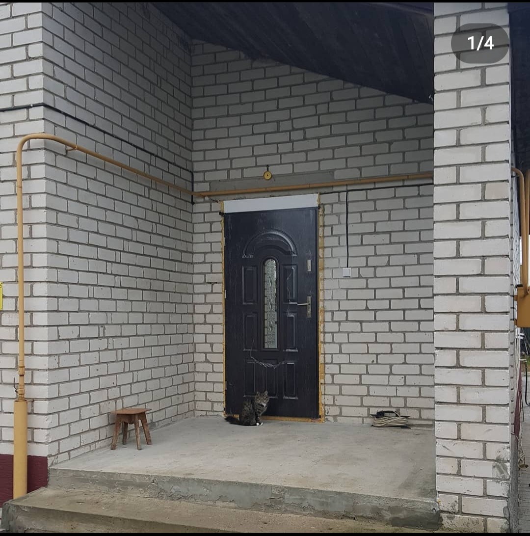 Wejście do domu z białej cegły z czarnymi drzwiami, gdzie siedzi kot. Widoczne rury gazowe i drewniany stołek.