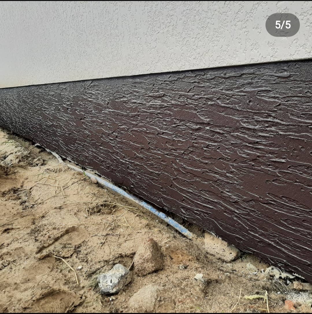 Fragment elewacji budynku z wyraźną, poziomą fakturą w odcieniach brązu, oddzielony od białej ściany cienką linią. Przy ziemi widoczny fragment niebieskiej rurki oraz piasek z kamieniami.