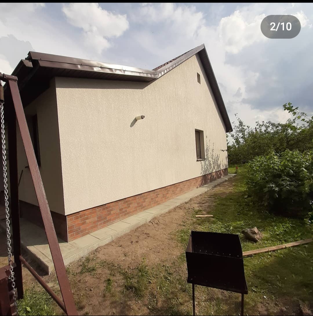 Jasna elewacja domu z ceglanym cokołem i brązowym dachem, widok z boku na tle ogrodu z grillem.