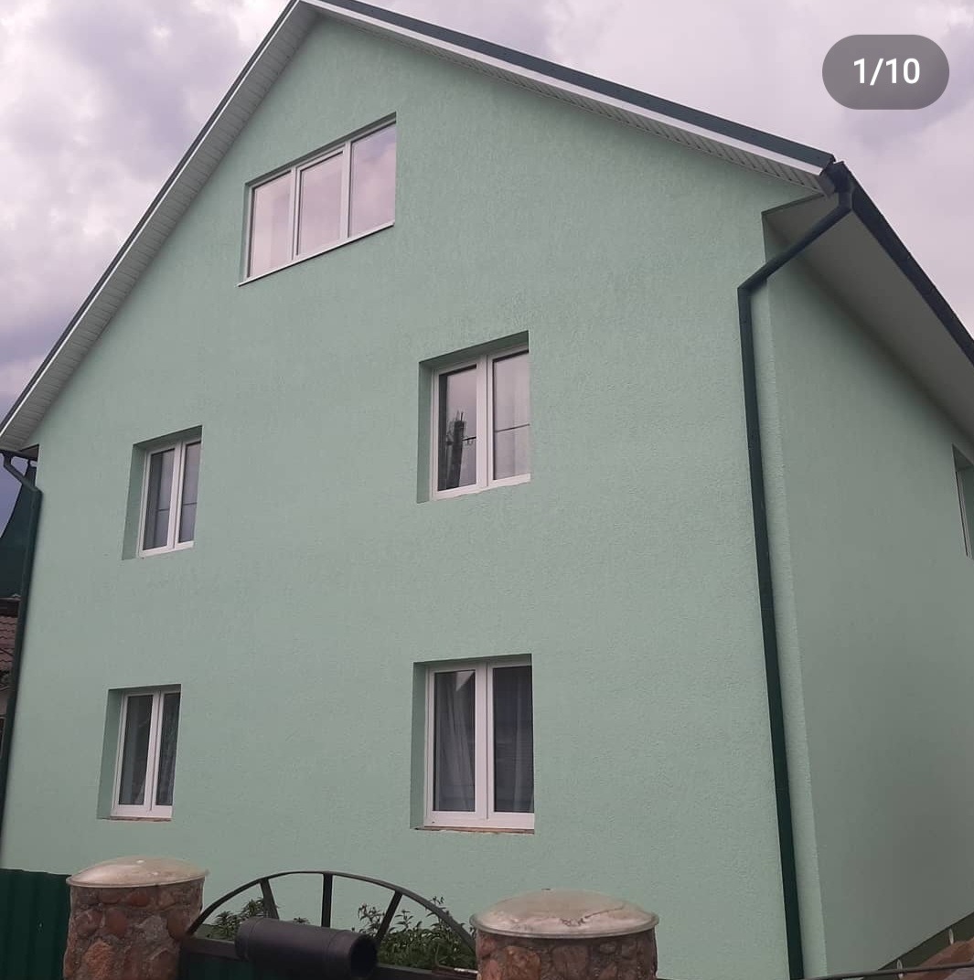 Dwukondygnacyjny dom z nową, jasną, pistacjową elewacją o strukturze baranka, białe ramy okienne, widok z dołu, fragment ogrodzenia z kamiennymi słupkami na pierwszym planie.