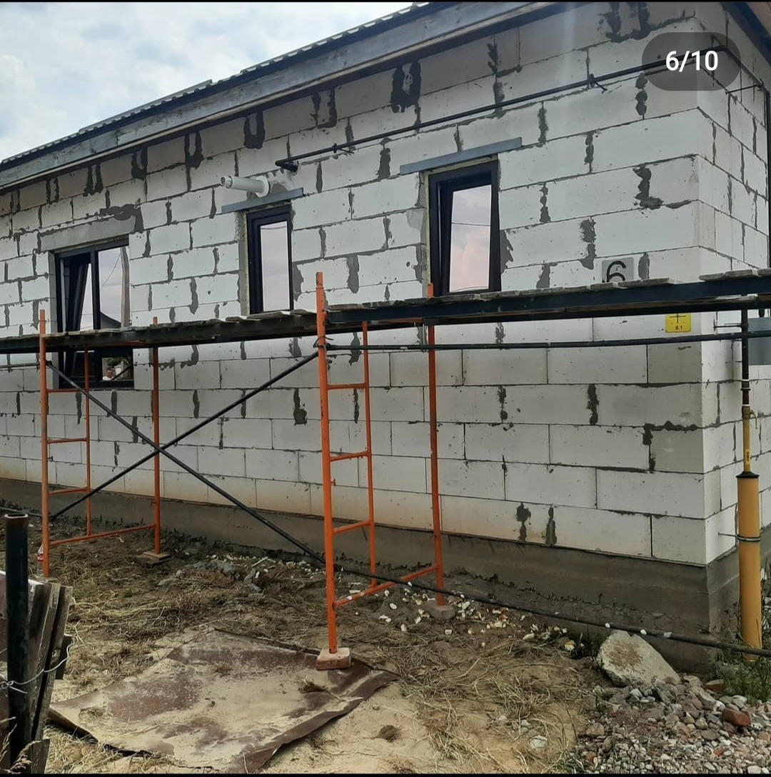 Budynek w trakcie wznoszenia ścian z bloczków betonowych, widoczne rusztowanie i okna z ciemnymi ramami.