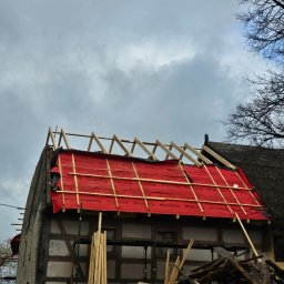 Ogólnobudowlane Rafal Stanislawski - Dach domu w trakcie wymiany, zabezpieczony czerwoną membraną i drewnianą konstrukcją. Widoczne stare elementy dachu i fragmenty drzew. Pochmurne niebo w tle.