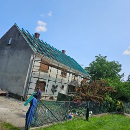 Ogólnobudowlane Rafal Stanislawski - Dom w trakcie wymiany dachu. Widoczne rusztowania, niebieskie elementy konstrukcji dachu i pracownik w trakcie pracy. Ujęcie z dołu, słoneczny dzień.
