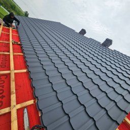 Ogólnobudowlane Rafal Stanislawski - Montaż grafitowej blachodachówki modułowej na dachu z widoczną membraną i łatami. W tle pochmurne niebo i zieleń. Na pierwszym planie piła spalinowa.