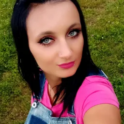 Portret kobiety z ciemnymi włosami i niebieskimi oczami, ubrana w różową koszulkę i dżinsowy kombinezon, na tle zielonej trawy.