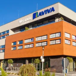 Nowoczesny budynek biurowy z pomarańczową elewacją i licznymi oknami, na dachu widoczne logotypy firm, w tle błękitne niebo.