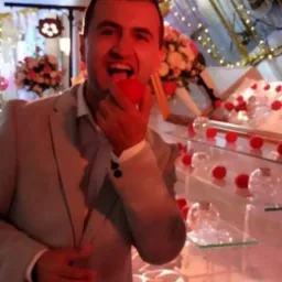 Uśmiechnięty mężczyzna w jasnym garniturze, z otwartymi ustami, trzyma w dłoni czerwoną kulkę, prawdopodobnie owoc, na tle dekoracji z kwiatów i szklanych kul na półkach.