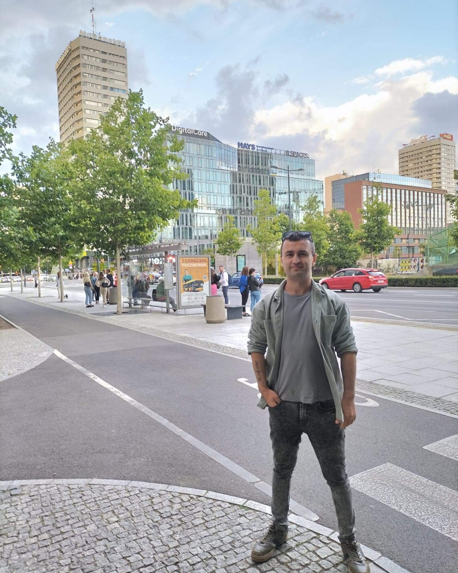 Mężczyzna w casualowym stroju stoi na tle miejskiego krajobrazu z nowoczesnymi biurowcami i przystankiem autobusowym.
