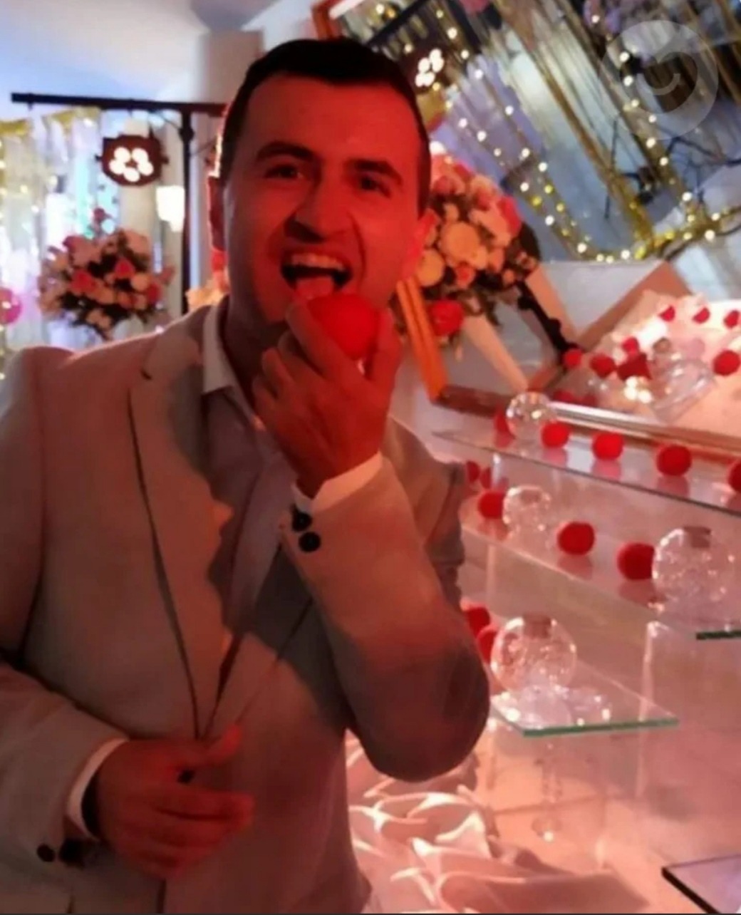 Uśmiechnięty mężczyzna w jasnym garniturze, z otwartymi ustami, trzyma w dłoni czerwoną kulkę, prawdopodobnie owoc, na tle dekoracji z kwiatów i szklanych kul na półkach.