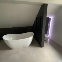 Nowoczesna łazienka na poddaszu z wolnostojącą białą wanną, czarną ścianką działową i grzejnikiem drabinkowym oraz toaletą w tle, wykończona jasnymi płytkami podłogowymi.