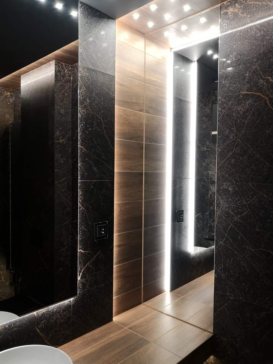 Elegancka łazienka wykończona ciemnym marmurem z wyraźnym użyleniem, podświetlane lustro z wbudowanymi punktowymi lampami sufitowymi, brązowe płytki podłogowe imitujące drewno.