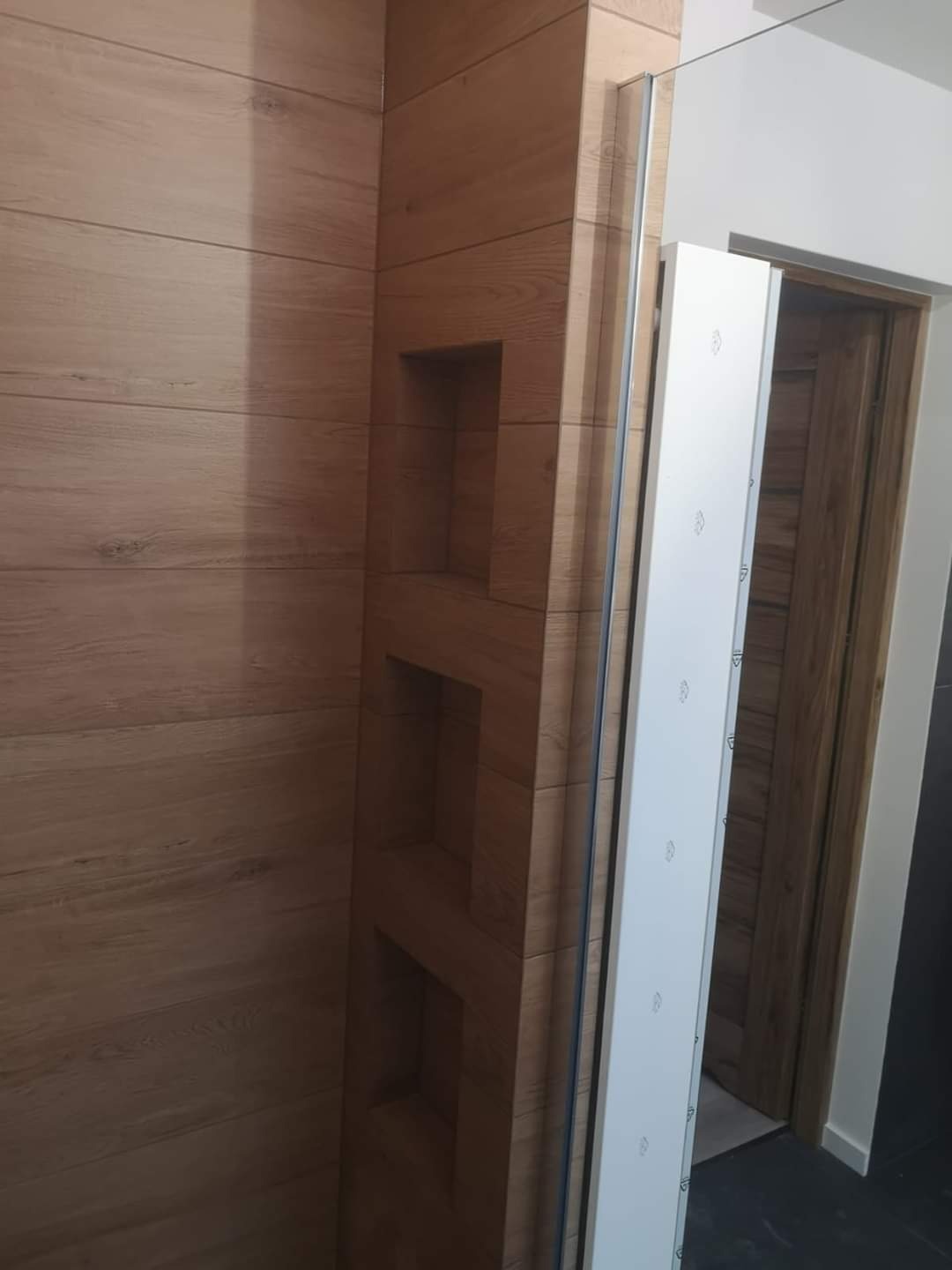 Wykończona ściana w łazience z płytkami imitującymi drewno, wbudowane półki, widoczna kabina prysznicowa i drzwi do łazienki.