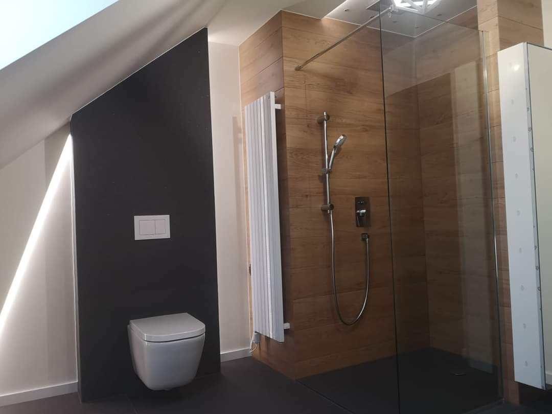 Nowoczesna łazienka na poddaszu z kabiną prysznicową typu walk-in z drewnopodobnymi płytkami, wiszącą toaletą na tle czarnej ściany i białym grzejnikiem drabinkowym.